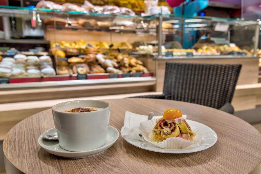 EDEKA-Abroell-Bad-Groenenbach-Baeckerei Tasse Kaffee und kleines Gebäck auf einem Tisch, im Hintergrund eine Café-Theke.